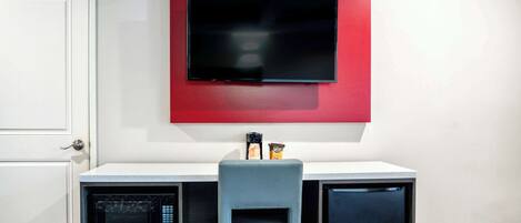 A compact room with a desk, a chair, a microwave, and a television mounted on the wall.