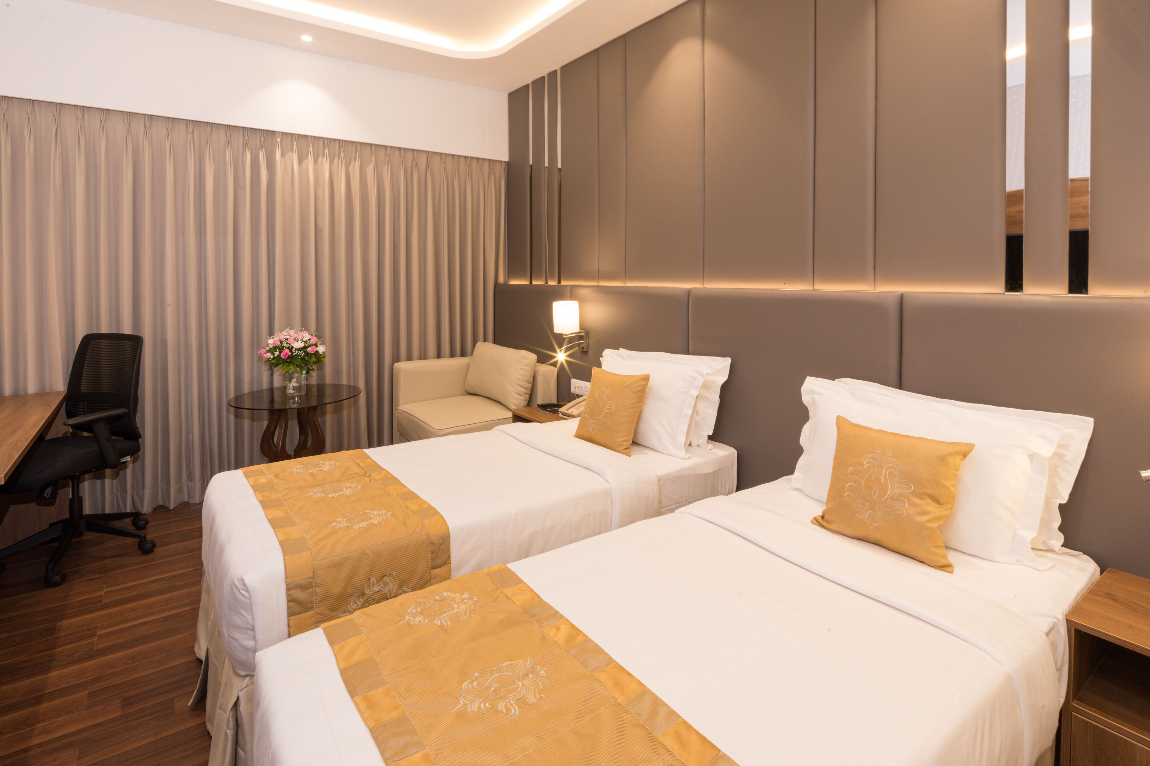 A hotel room with two beds, a desk, a chair, and a small table with a vase of flowers.
