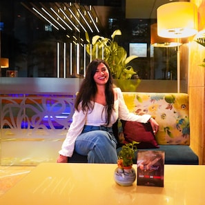 A woman sitting on a sofa in a well-lit lobby area with a modern design, including a decorative wall and a potted plant.