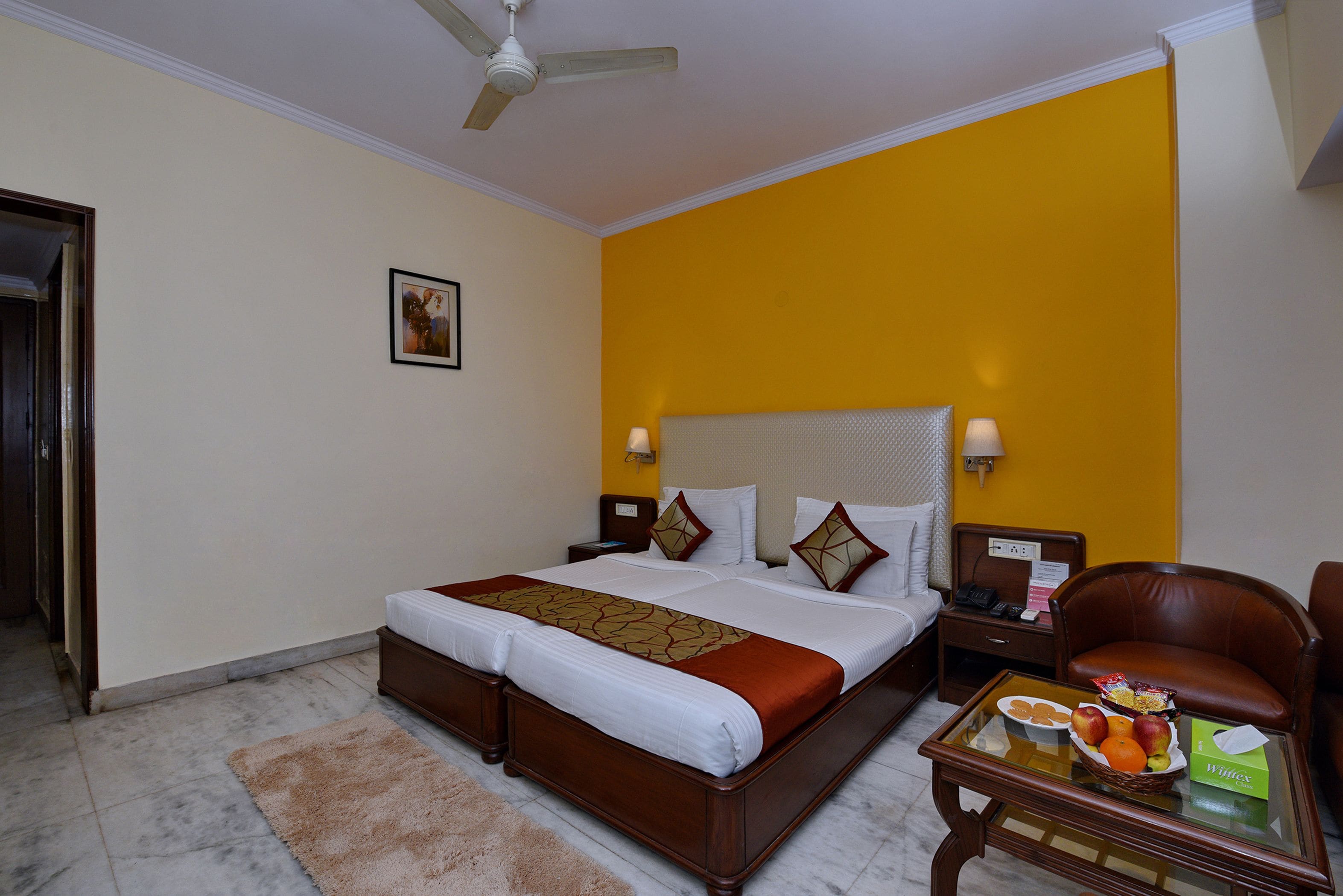 A hotel room with a bed, a nightstand, a chair, and a table with fruit and snacks.