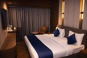 A hotel room with a large bed, a wooden desk, a chair, and a small table with a lamp. - The Beaufort Inn (New Delhi)