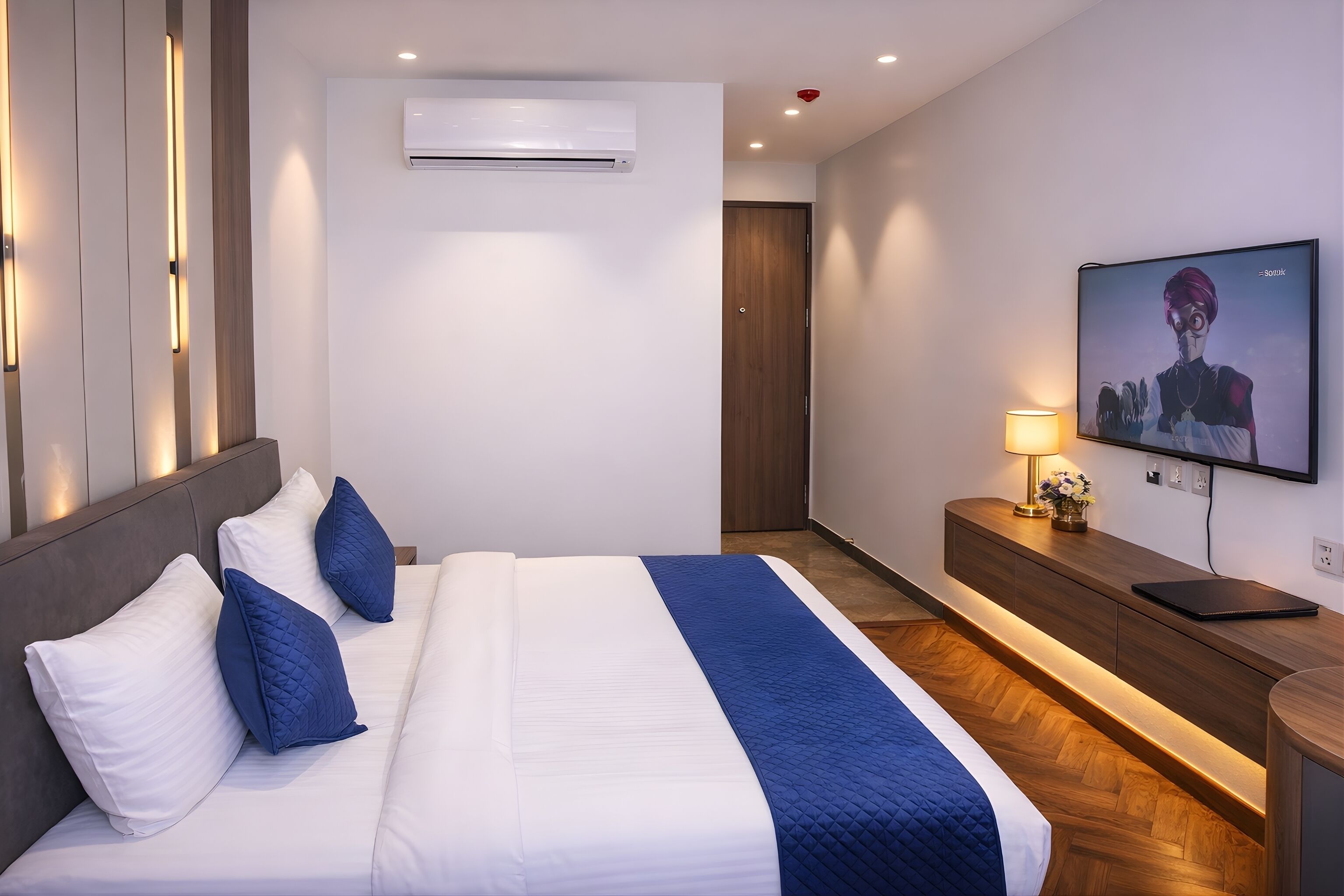 A hotel room with a bed, a TV, a bedside table with a lamp, and an air conditioning unit.