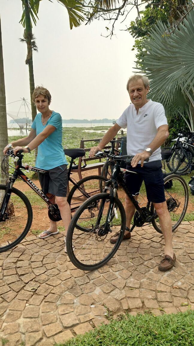 Two people standing next to bicycles on a paved path with a palm tree and a body of water in the background.