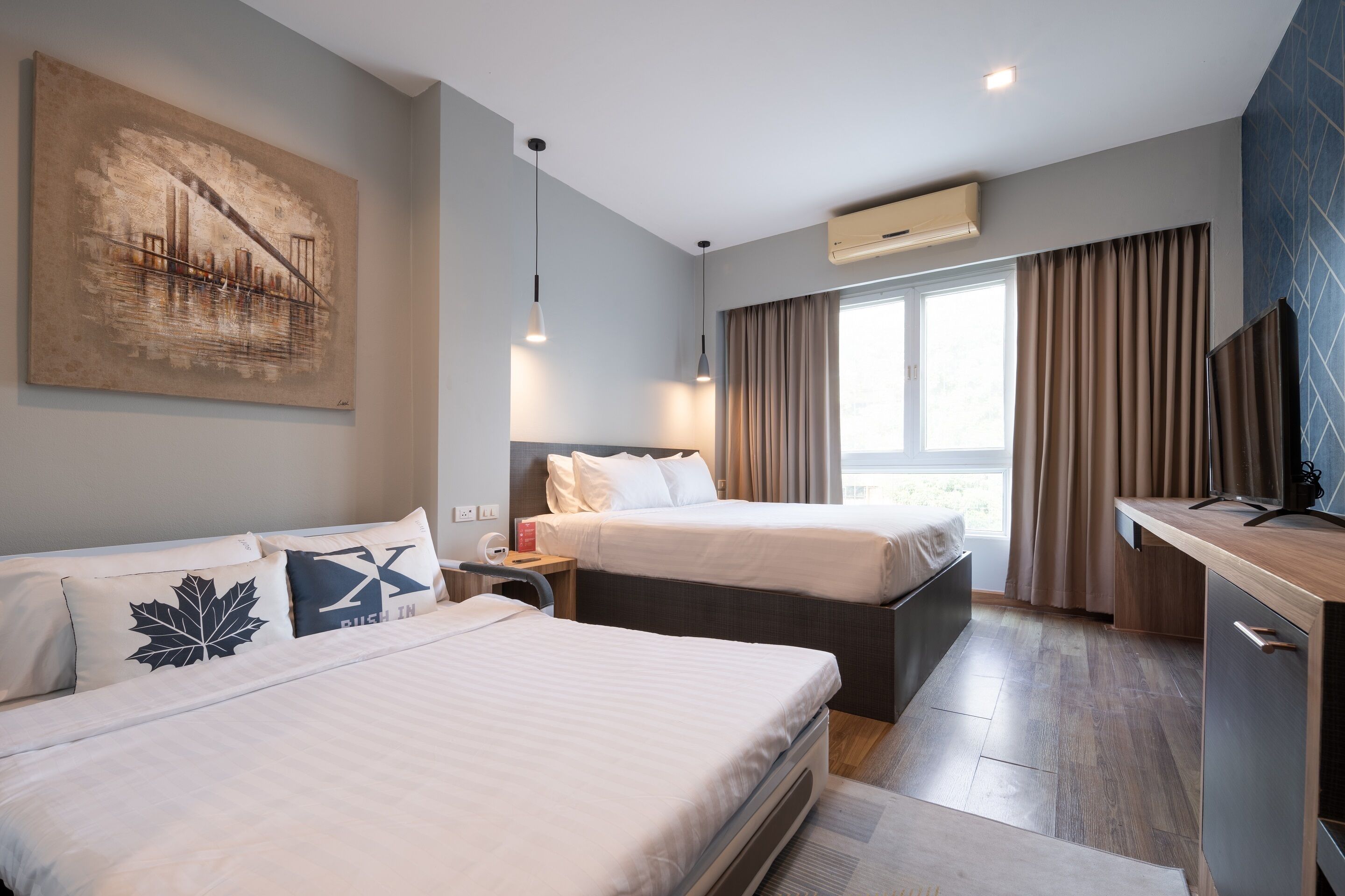 A hotel room with two beds, a desk, a large window with curtains, and an air conditioning unit.