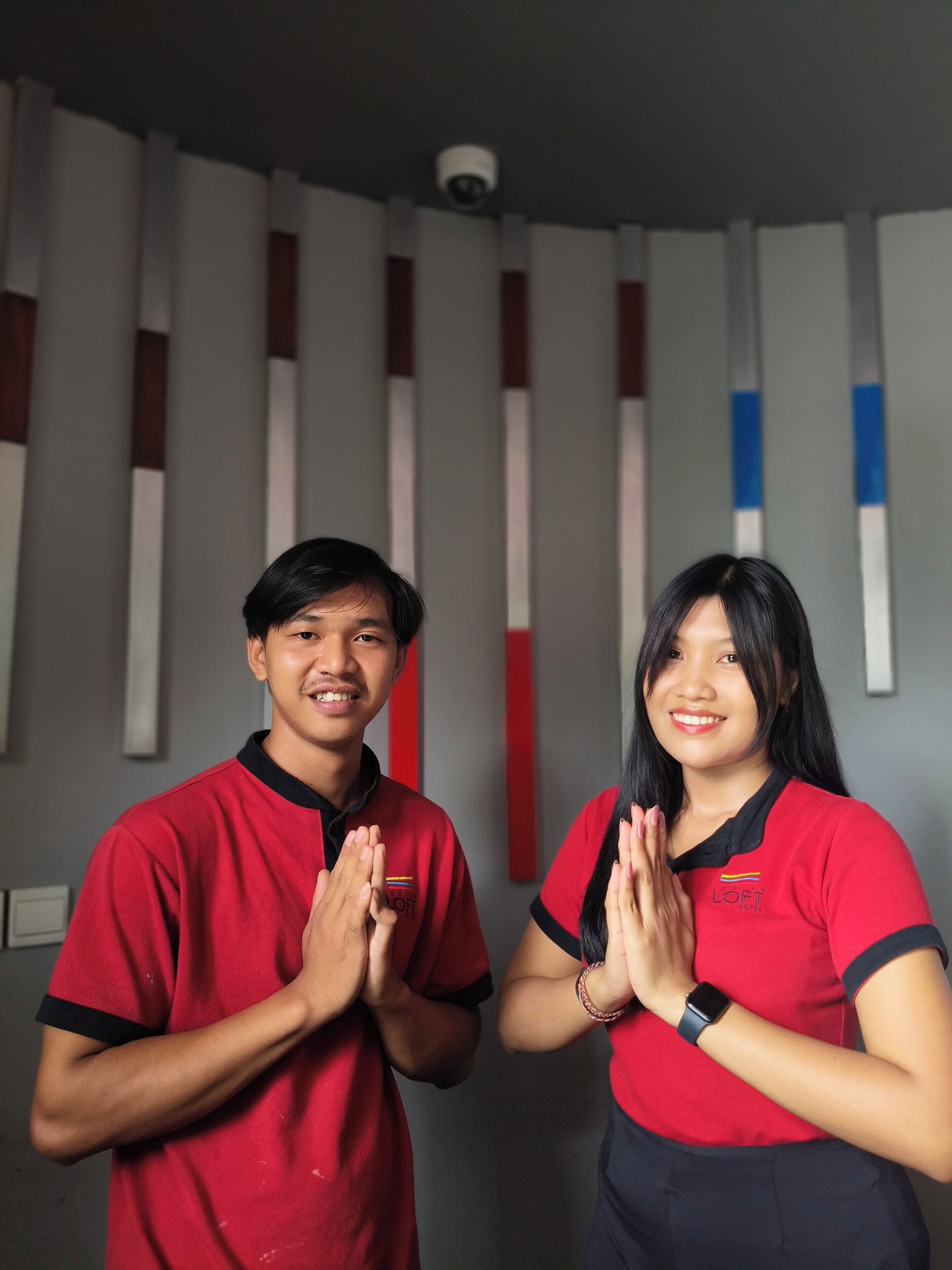Dos personas con camisetas rojas con un logotipo, de pie en una habitación con rayas verticales en la pared.