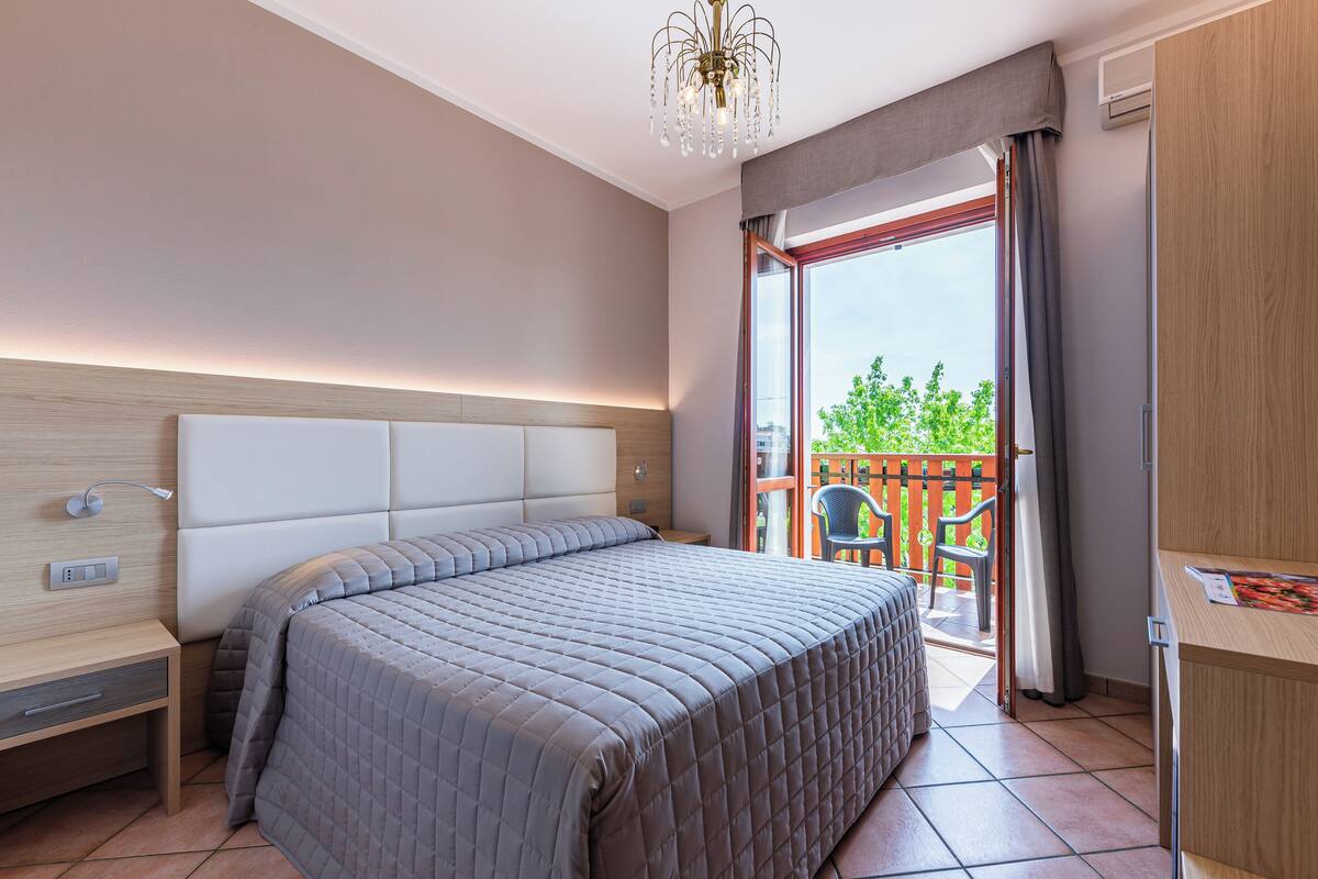 A modern bedroom with a large bed, bedside table, and a view of the outdoor balcony.