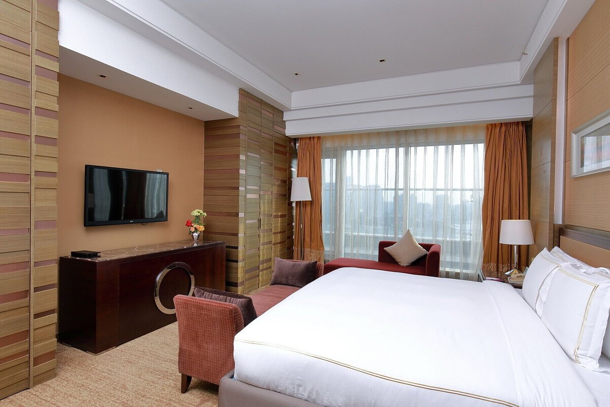 Ein Hotelzimmer mit einem großen Bett, einem an der Wand befestigten Fernseher, einem Schreibtisch mit Lampe und Blick auf die Stadt durch das Fenster.