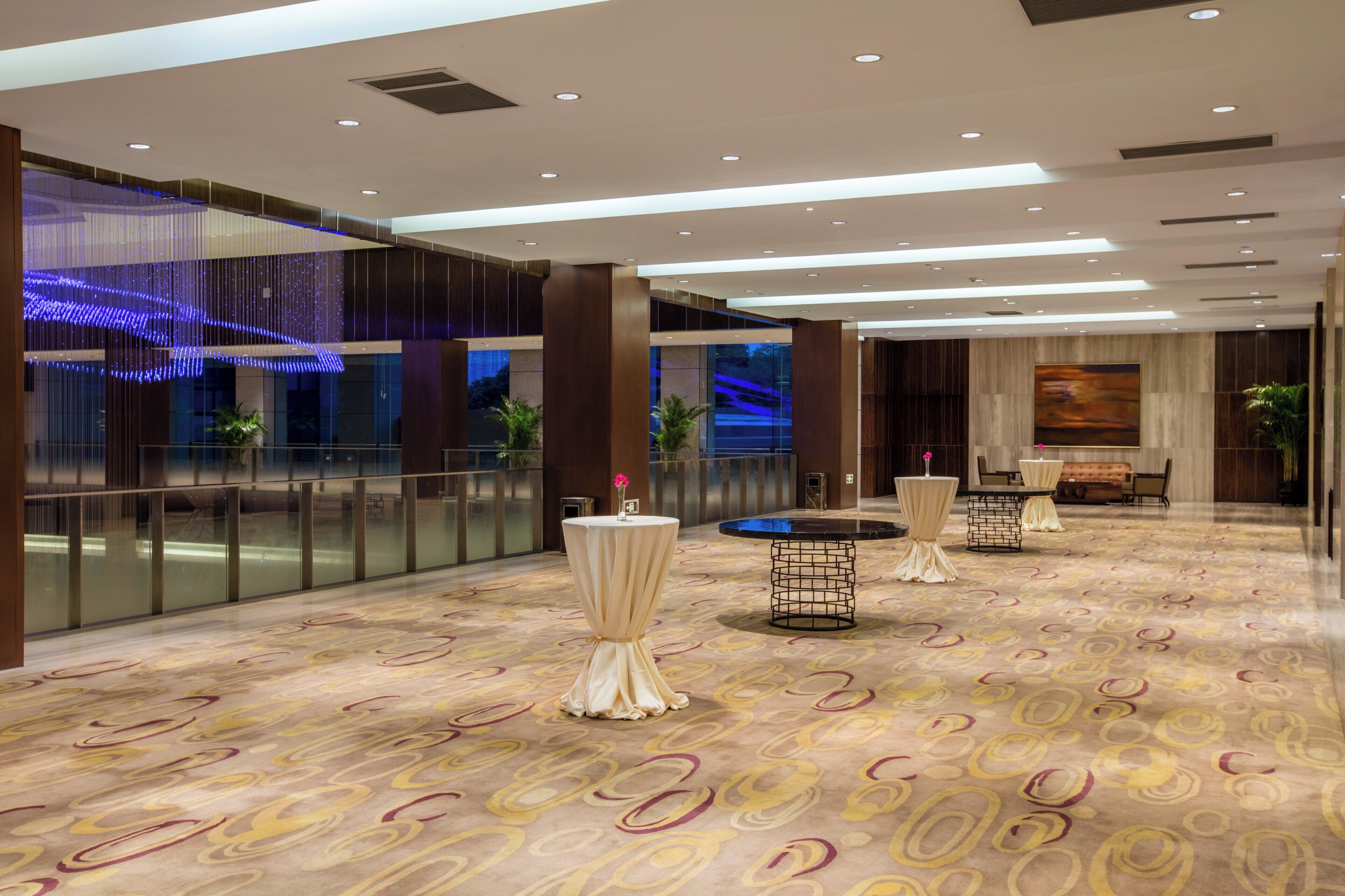 Un hall d’hôtel spacieux, doté d’un tapis à motifs, de tables rondes recouvertes de nappes blanches et d’un design de plafond moderne.