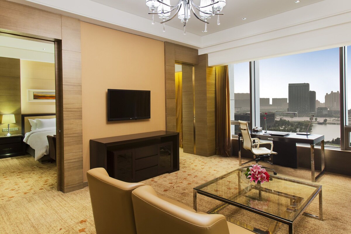 Ein modernes Hotelzimmer mit großem Fenster und Blick auf die Stadt, einem an der Wand befestigten Flachbildfernseher, einem Glastisch mit Blumenvase und einer gemütlichen Sitzecke mit Sofa und Sesseln.