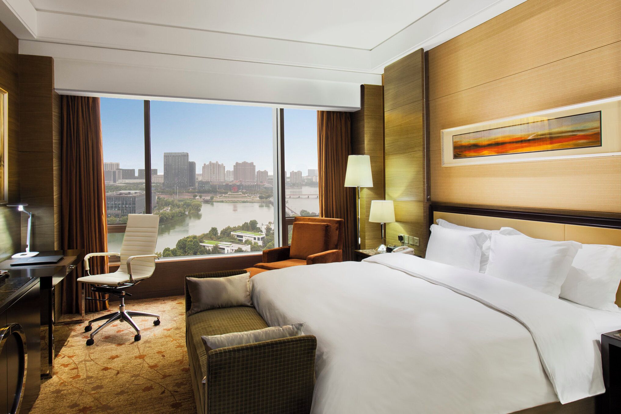 A hotel room with a large bed, a desk, a chair, a lamp, and a view of a cityscape.