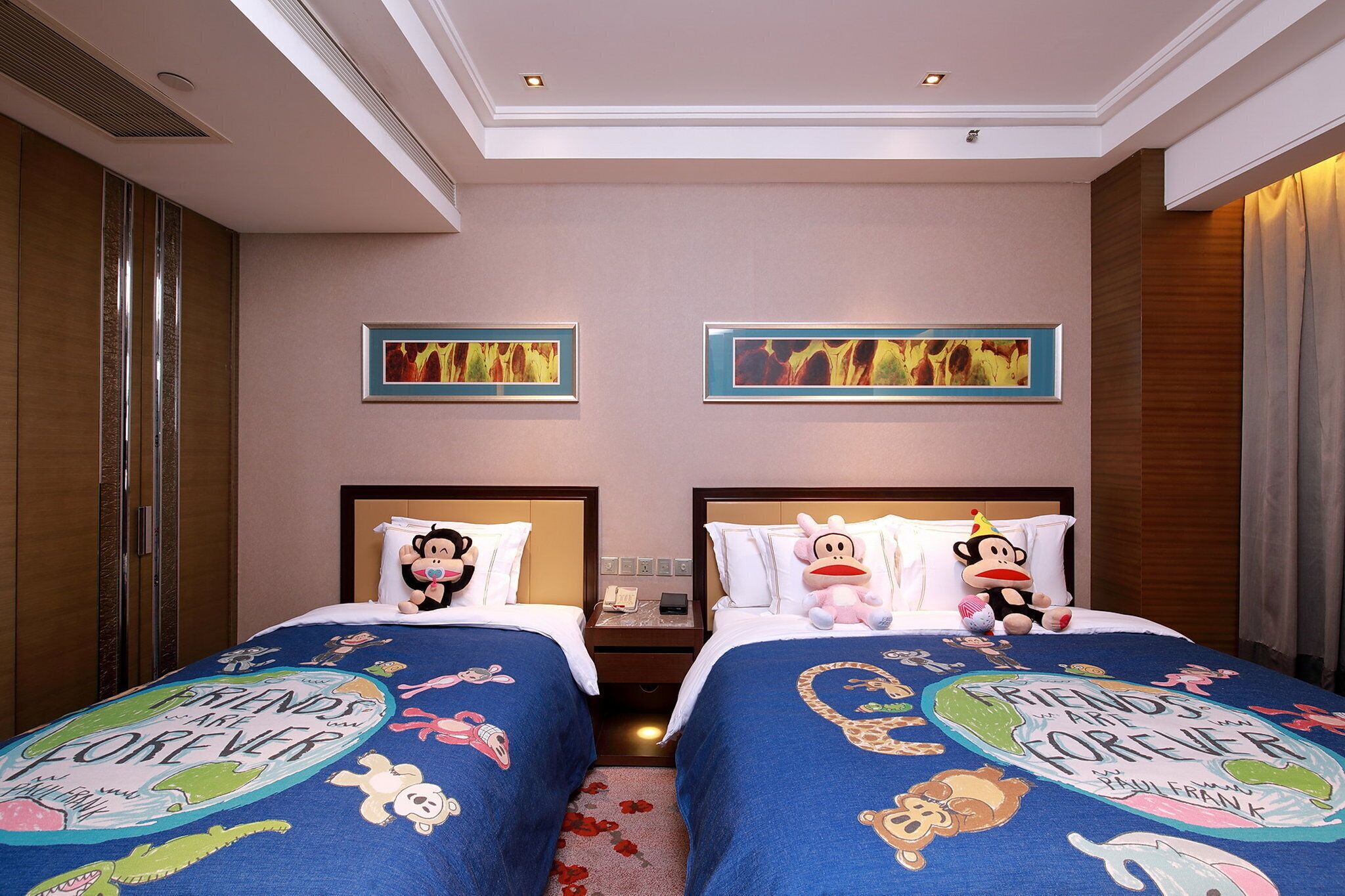 A hotel room with two beds, each with a blue bedspread featuring cartoon characters and the text 'Friends Forever.' There are two framed abstract paintings on the wall, a wooden wardrobe, and a nightstand with a phone and a lamp.