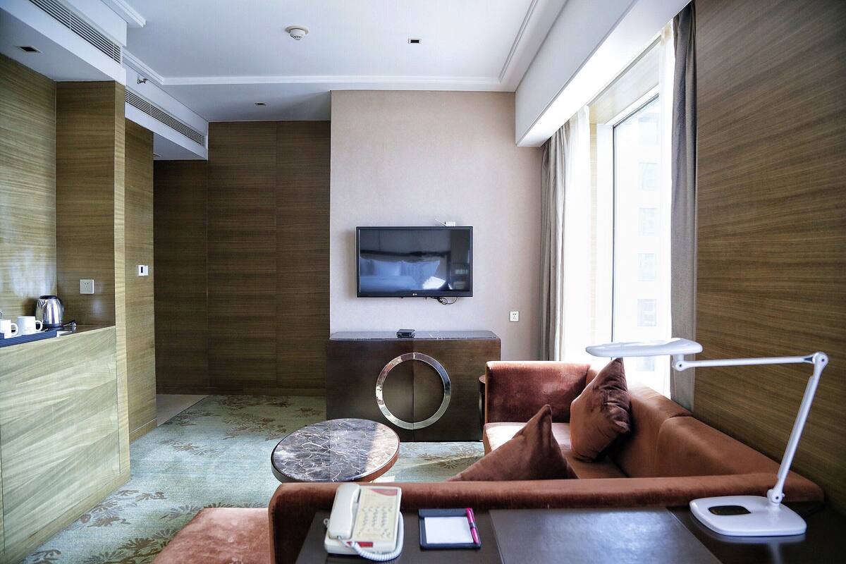 Una habitación de hotel moderna con sofá, una mesita, una lámpara de escritorio, televisión y minibar.