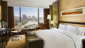 Een hotelkamer met een groot bed, een bureau, een stoel en uitzicht op een stadsgezicht door grote ramen.