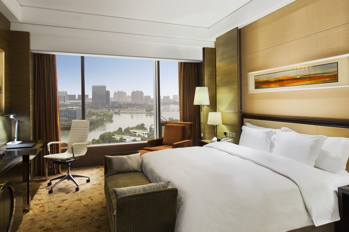 Ein Hotelzimmer mit einem großen Bett, einem Schreibtisch, einem Sessel und Blick auf eine Stadtlandschaft durch große Fenster.