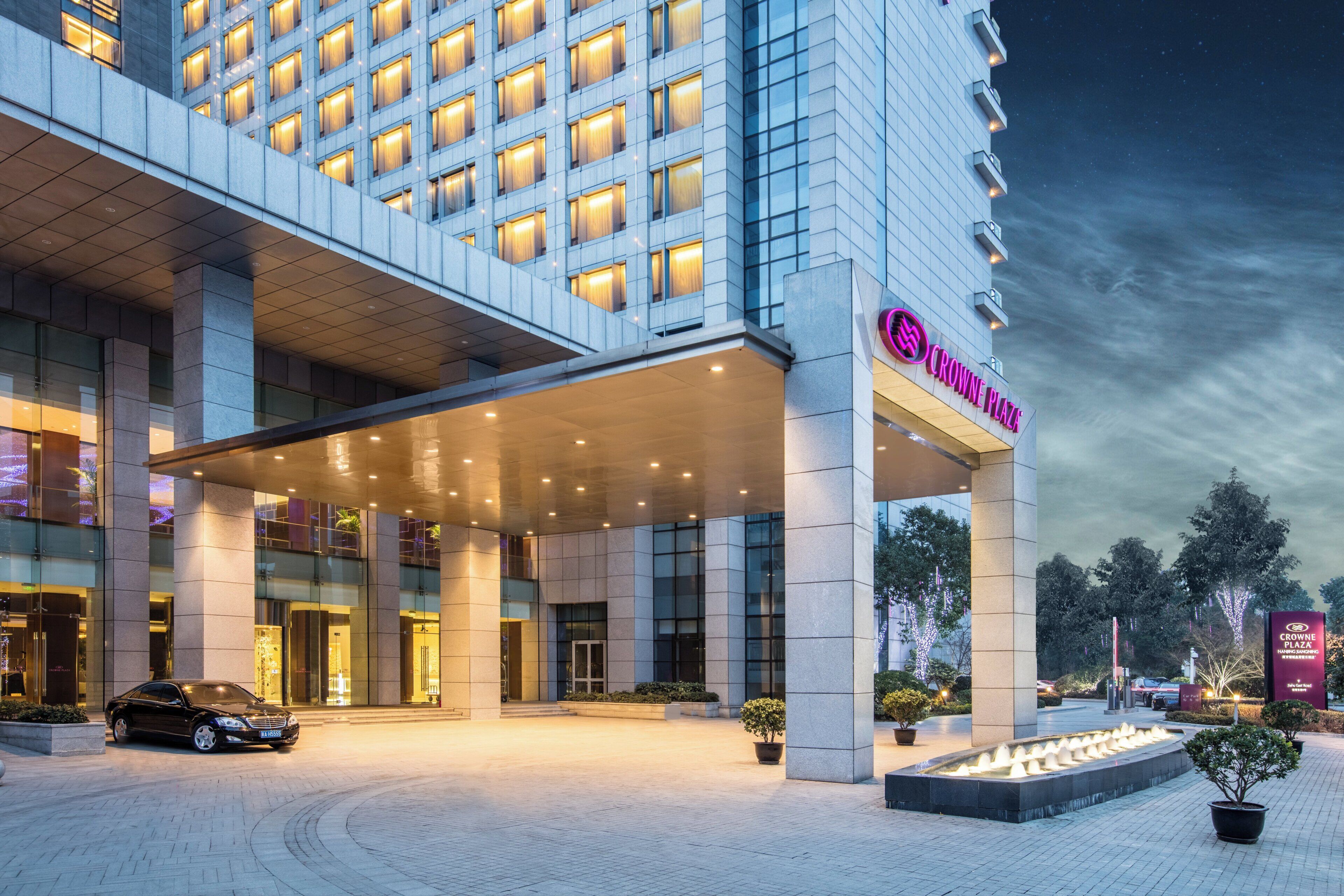Entrada de hotel moderna com hall de entrada coberto, um carro estacionado em frente e fachada bem iluminada.