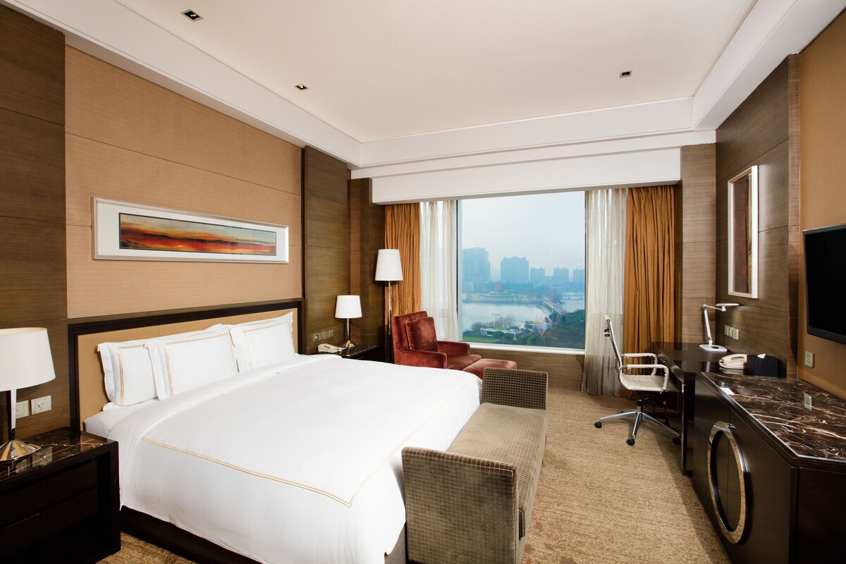 Ein Hotelzimmer mit einem großen Bett, einem Schreibtisch mit Stuhl, einem Fernseher und Blick auf die Stadt.