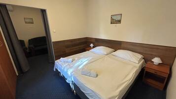 Une chambre d’hôtel avec un lit double, deux tables de chevet, une petite lampe de chevet, un tableau au mur et une porte donnant sur une autre pièce.