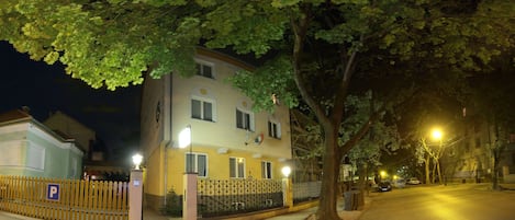 A multi-story building with a gated entrance, a parking sign, and a street with trees and streetlights.