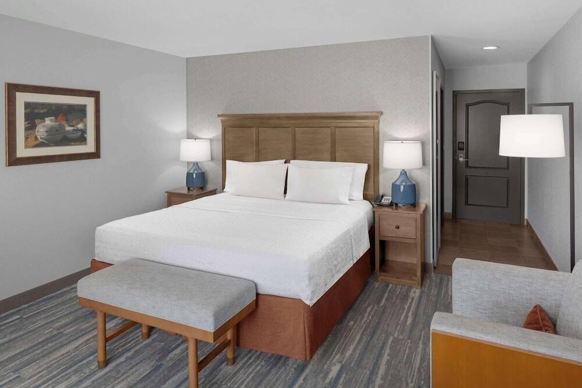 Habitación de hotel con cama, mesitas de noche, un banco, una silla, una puerta y un cuadro en la pared.