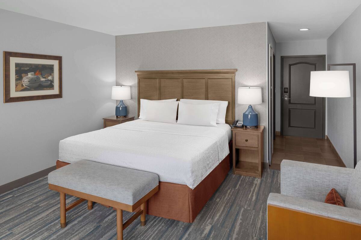 Habitación de hotel con una cama grande, dos mesitas de noche con lámparas, un banco y una puerta que da a un pasillo.