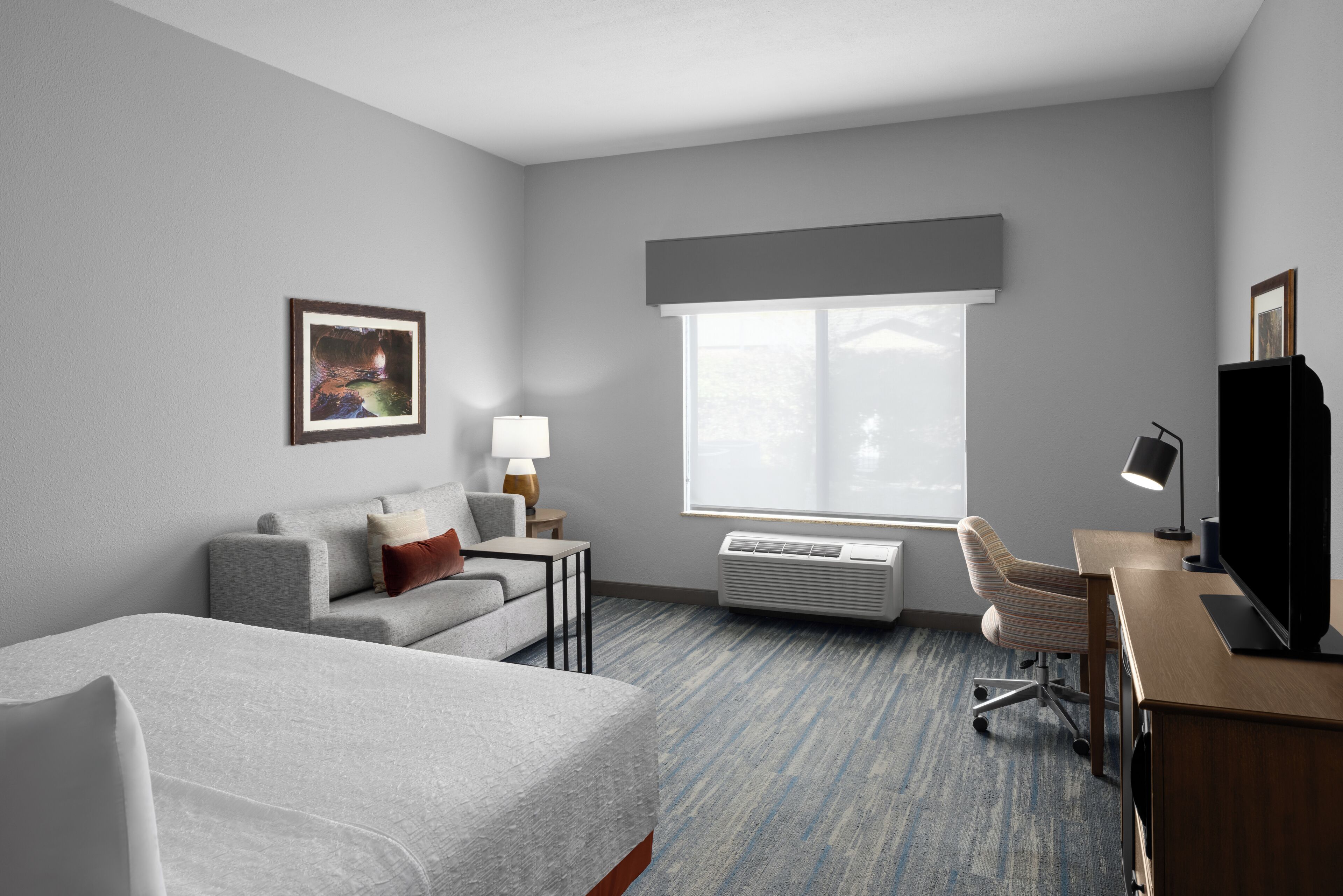 A hotel room with a bed, a sofa, a desk with a computer, a TV, a window, and an air conditioner.