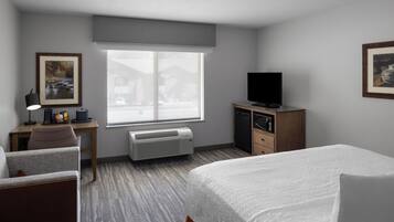 Une chambre d’hôtel comprenant un lit, un bureau avec une chaise, une télévision, un radiateur et une fenêtre donnant sur des immeubles.