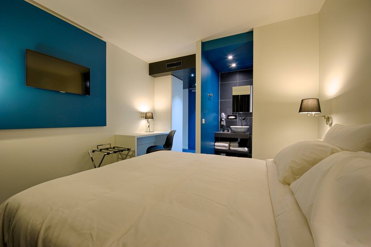 A hotel room with a bed, desk, chair, and a bathroom with a mirror and sink.