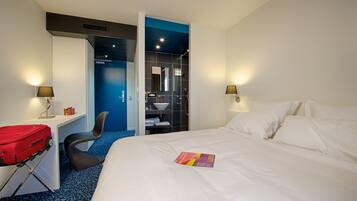A hotel room with a bed, a desk with a red suitcase, a chair, and a bathroom visible through an open door.