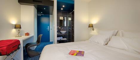 A hotel room with a bed, a desk with a red suitcase, a chair, and a bathroom visible through an open door.