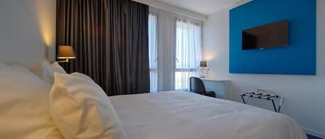 Habitación de hotel con una cama grande, un escritorio con silla, un televisor montado en la pared y una ventana con cortinas.