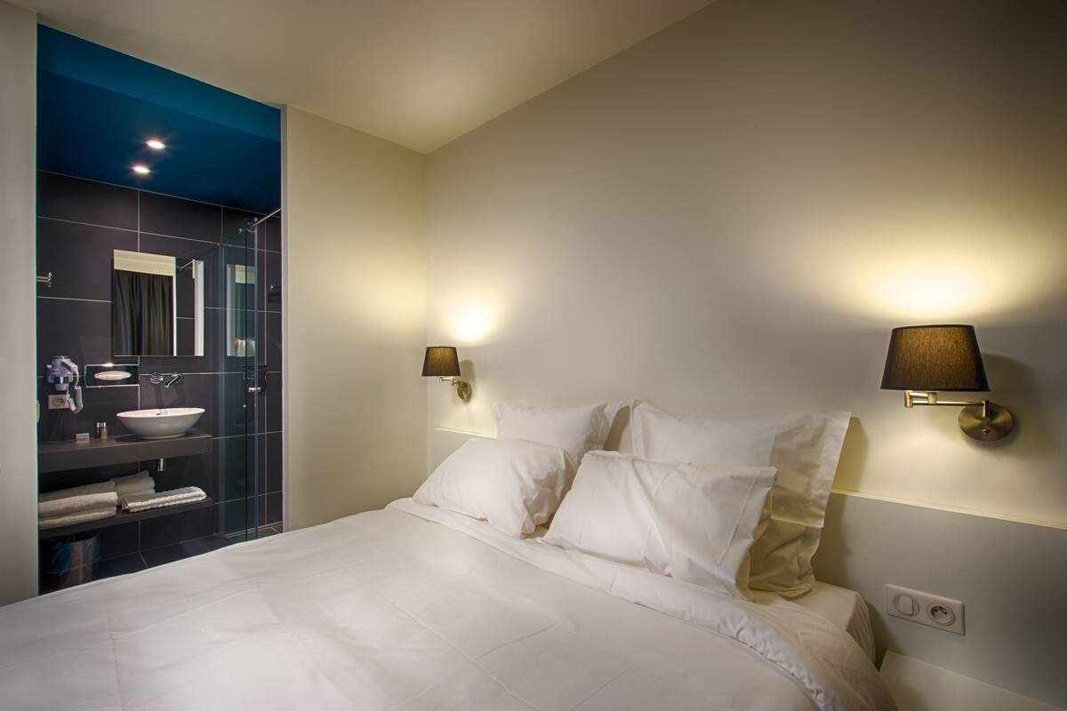 A modern hotel room with a large bed, a bathroom with a glass shower enclosure, and two wall-mounted lamps.