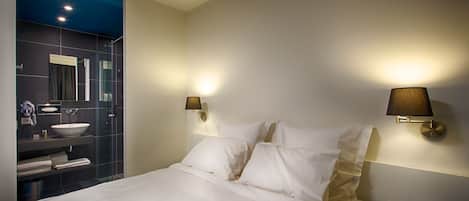 Quarto de hotel moderno com uma cama grande, banheiro com box de vidro e duas luminárias fixadas na parede.