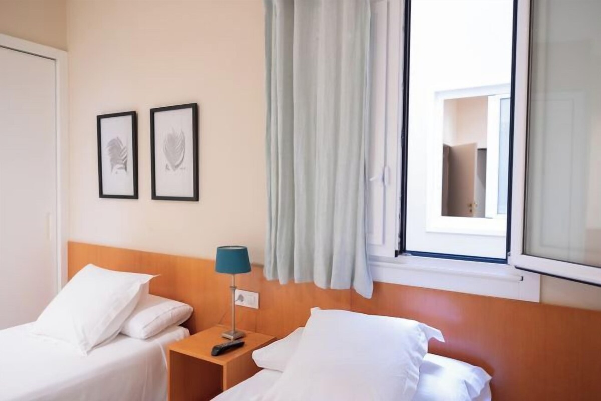 Habitación de hotel con dos camas, una mesita de noche, una lámpara y un ventanal con cortinas.
