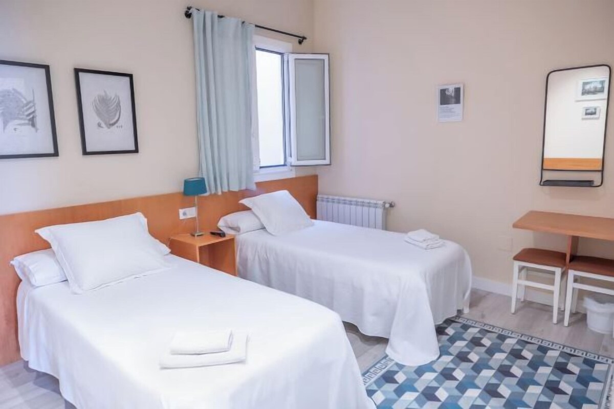 A hotel room with two beds, a desk, a chair, a radiator, and a window with curtains.