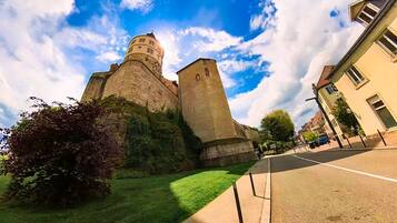 Eine historische Burg mit einem markanten runden Turm und einem kleineren rechteckigen Turm, umgeben von einer gepflegten Rasenfläche und einer Straße mit Gebäuden.