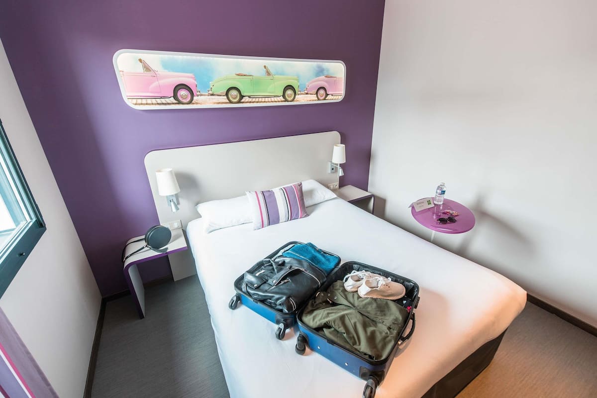 Habitación de hotel con cama, maleta, almohada, mesita de noche, lámpara, un cuadro con coches y una pequeña mesa con una botella de agua.