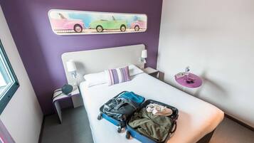 Une chambre d’hôtel avec un lit, une valise, un oreiller, une table de chevet, une lampe, une décoration murale représentant des voitures et une petite table avec une bouteille d’eau.
