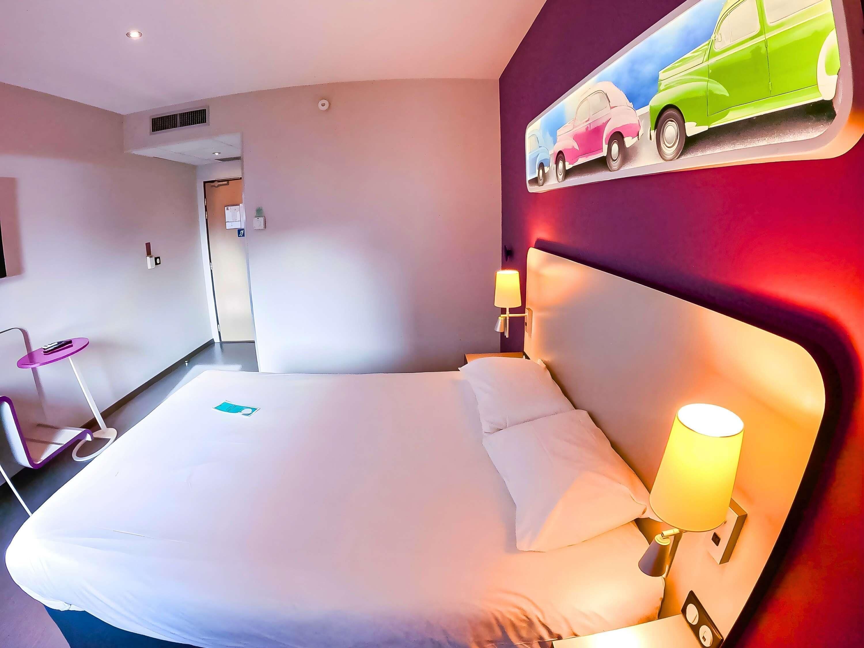 A hotel room with a large bed, a headboard with a light fixture, a wall-mounted painting of cars, and a small table with a chair.
