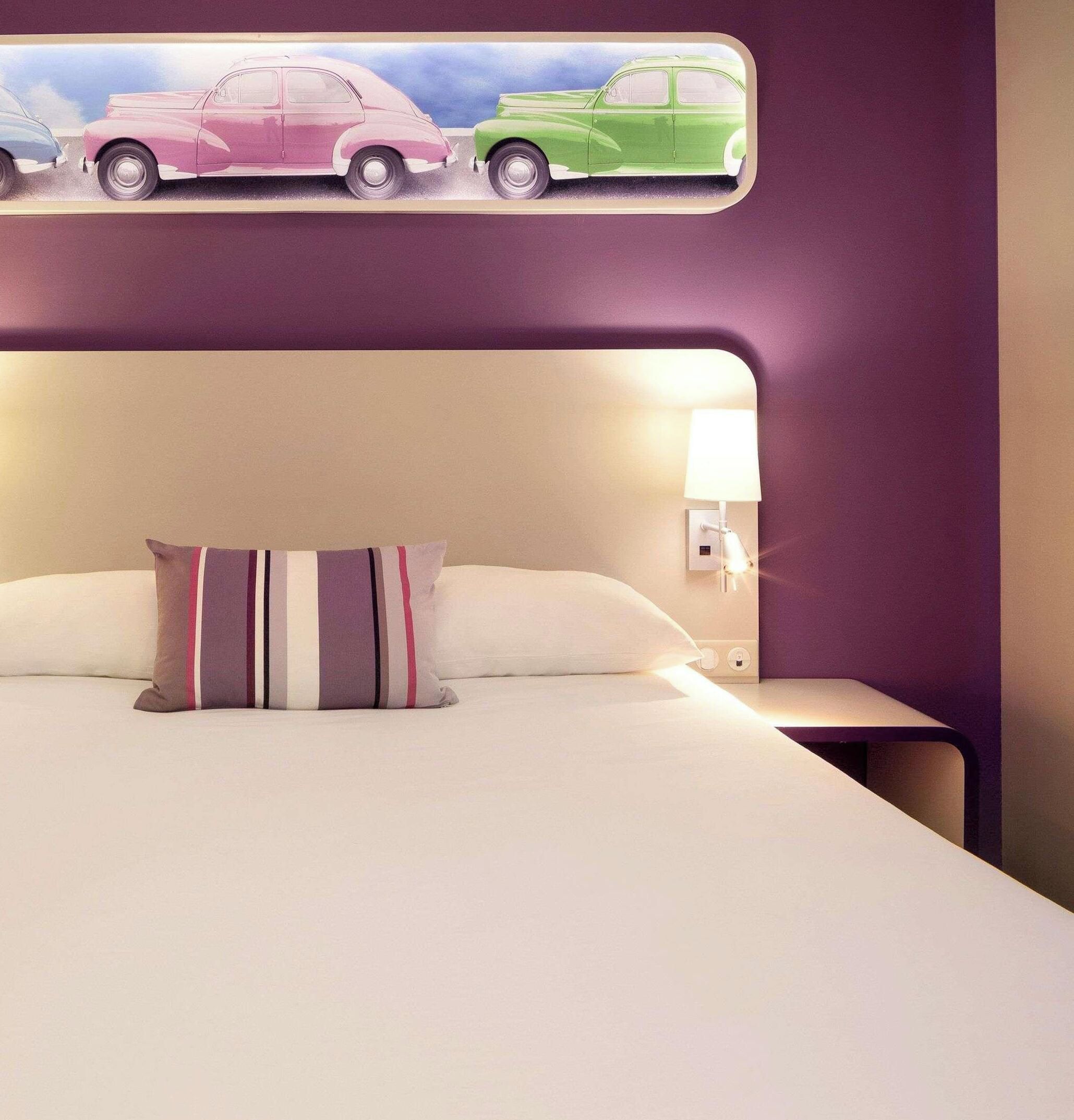Quarto de hotel com cama, almofadas, mesa de cabeceira, luminária fixada na parede e uma obra de arte retratando carros antigos.