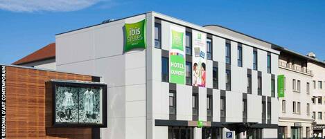 Un moderno edificio de hotel con varios paneles verdes con el logo 'ibis Styles', un letrero que dice 'Hotel Ouvert' y un letrero que indica 'Parking Velotte'.