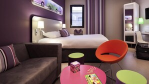Una habitación de hotel moderna con una cama grande, un sofá, una mesita redonda con un libro y una caja, y una silla naranja brillante.
