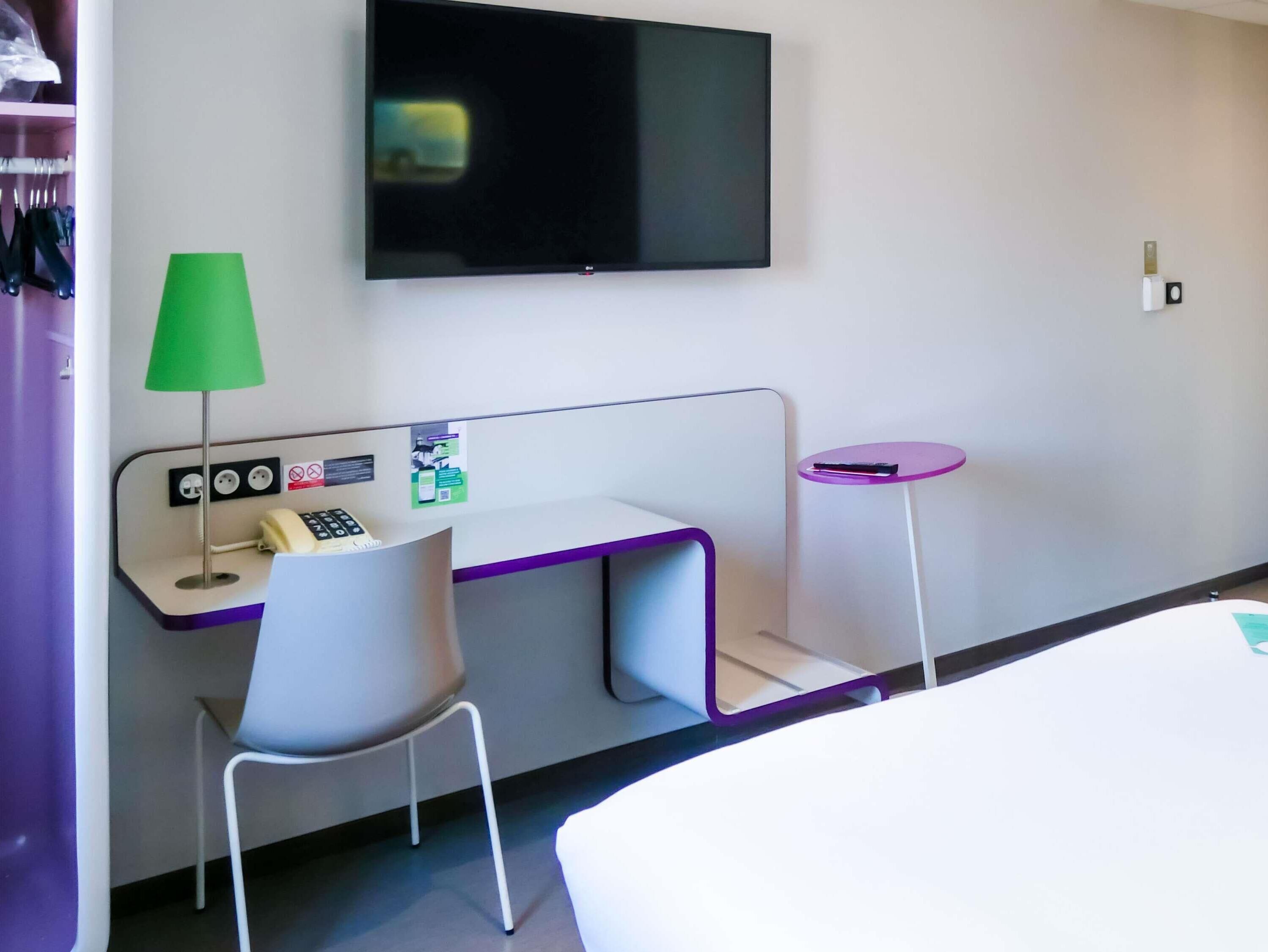 A hotel room with a desk, chair, TV, and a small table.