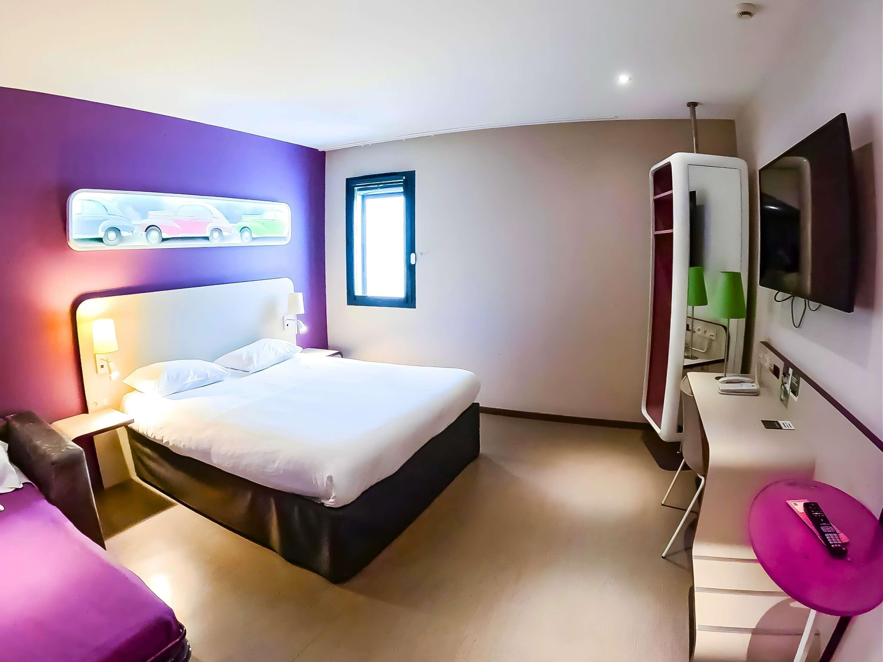 A hotel room with a bed, bedside table, lamp, TV, and a small table with a remote control.
