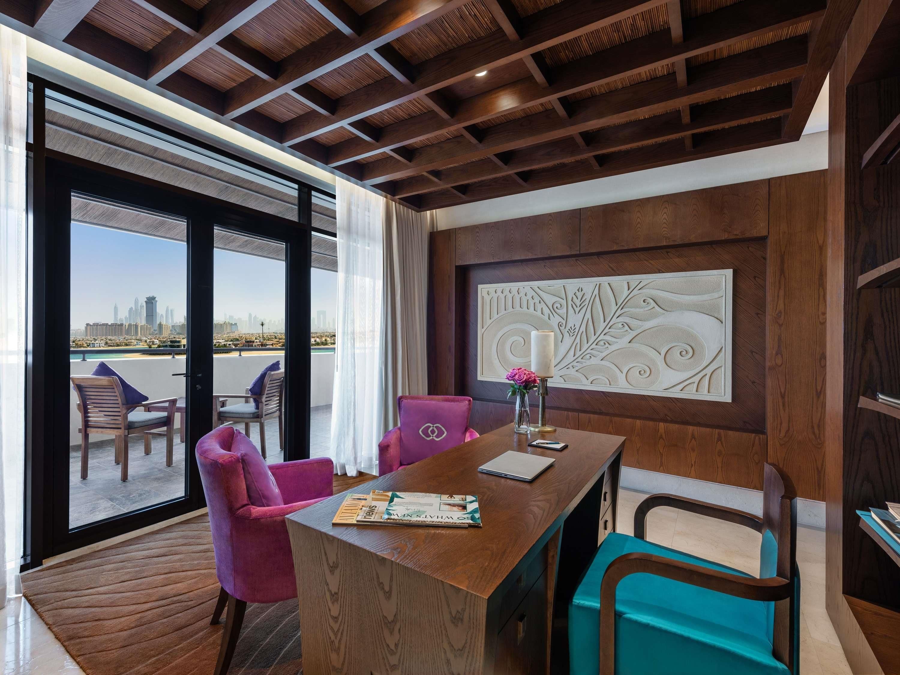 A modern hotel room with a wooden ceiling, a large window with a city view, a dining area with a wooden table, and a decorative wall panel.