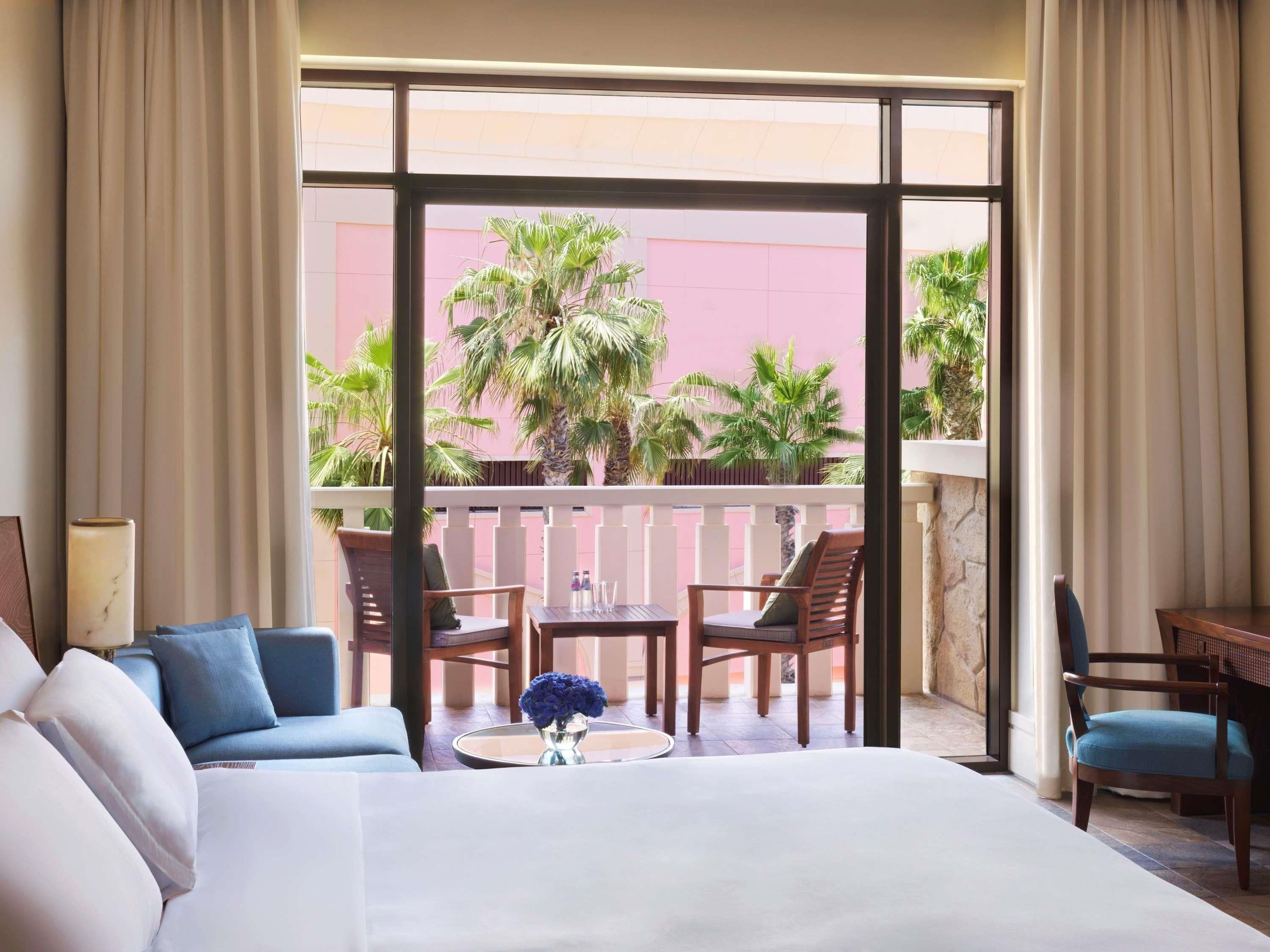 Ein Hotelzimmer mit einem großen Bett, einem Balkon mit Stühlen, einem kleinen Tisch und Blick auf Palmen.