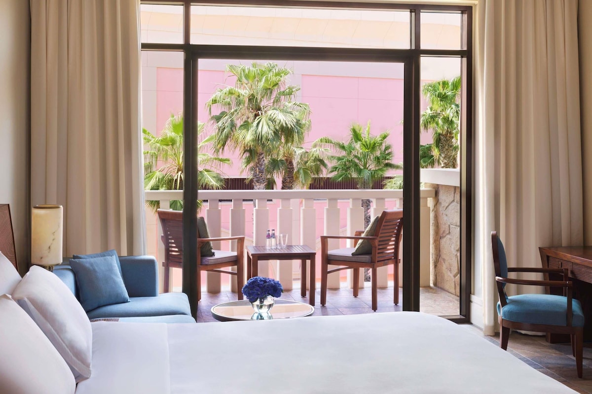 Habitación de hotel con una cama grande, un balcón con sillas, una mesita y vistas a palmeras.