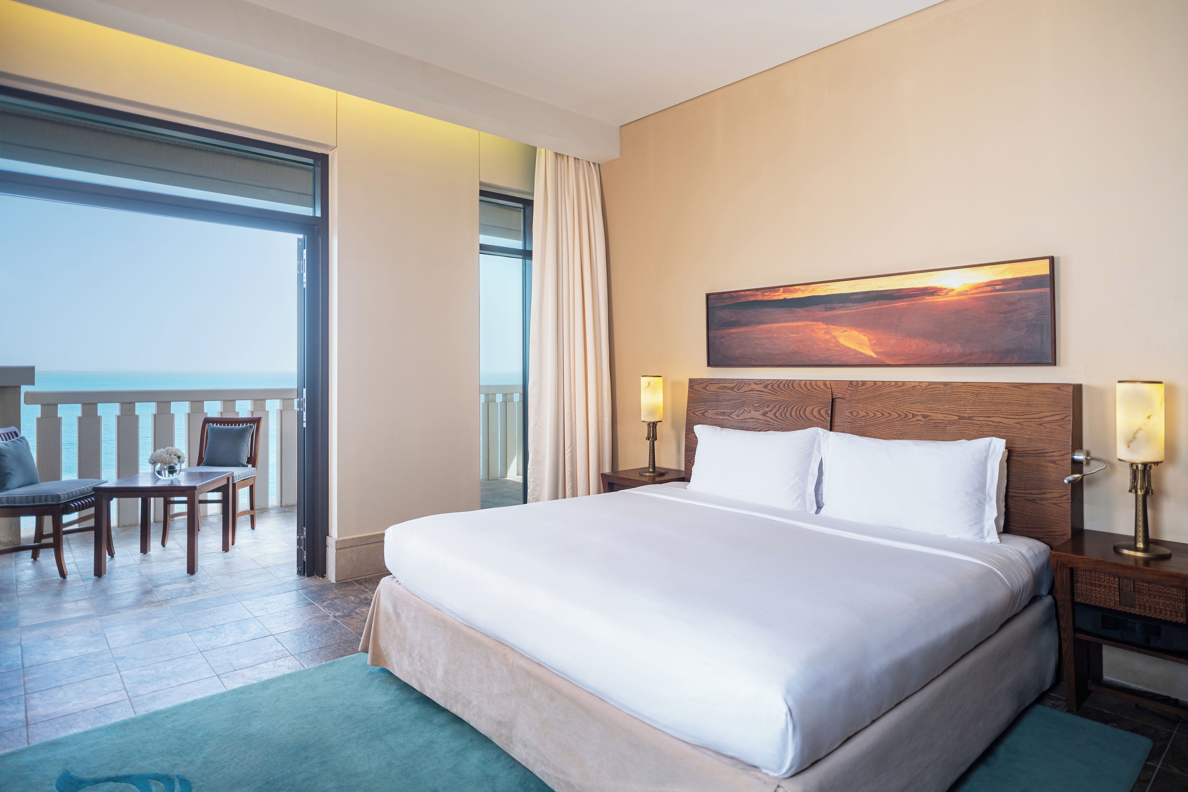 A hotel room with a large bed, a painting of a sunset, a desk with a chair, and a view of the sea through a balcony.