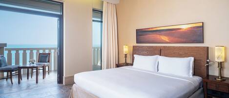 A hotel room with a large bed, a painting of a sunset, a desk with a chair, and a view of the sea through a balcony.