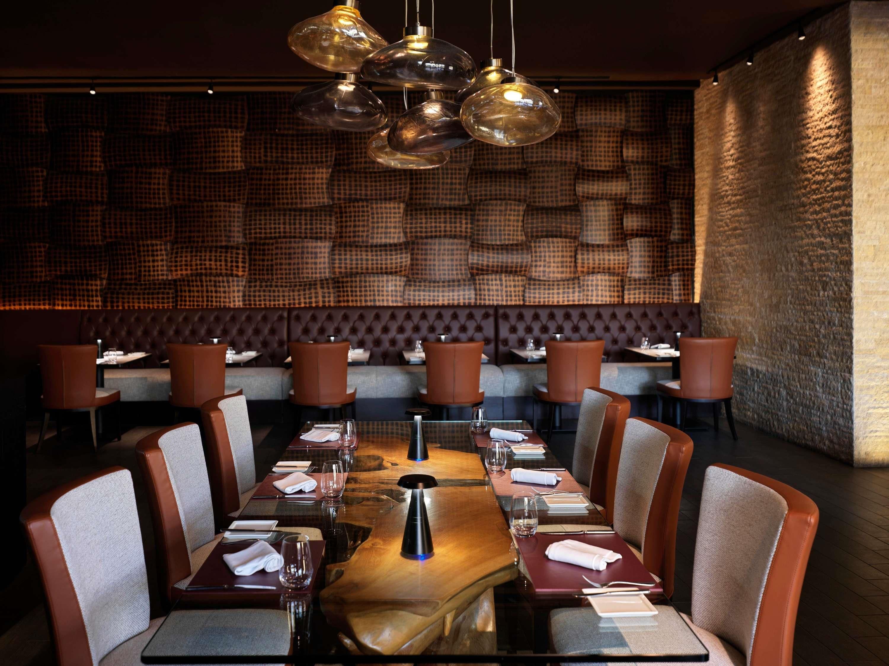 A restaurant with a long wooden table, brown upholstered chairs, and a patterned wall in the background.