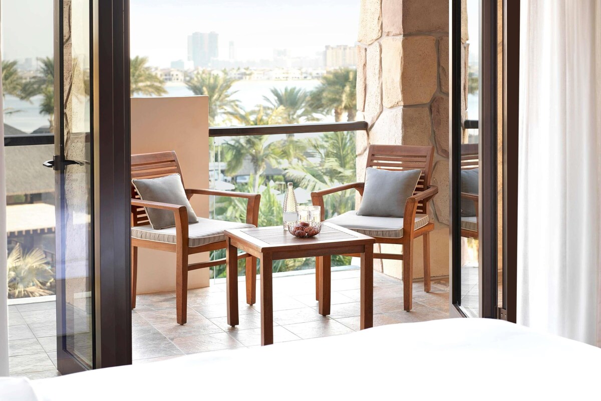 Een balkon met twee houten stoelen, een kleintje tafeltje en uitzicht op palmbomen en gebouwen.